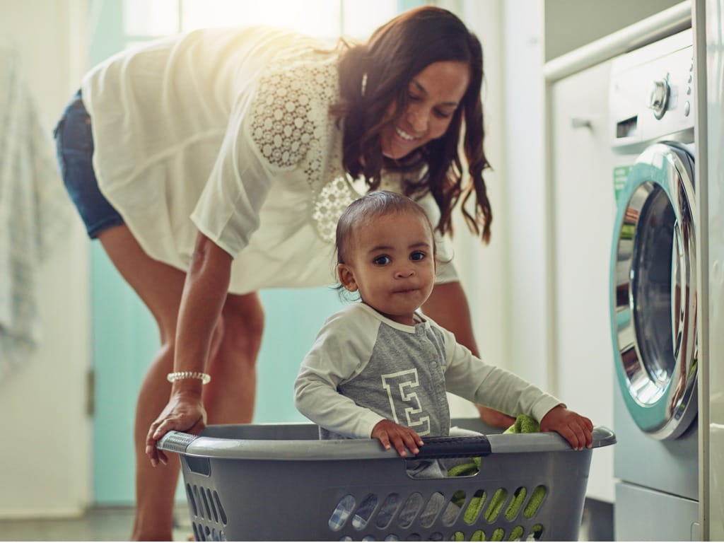 kid sitting in laundry basket next to mother putting clothes in the washing machine
